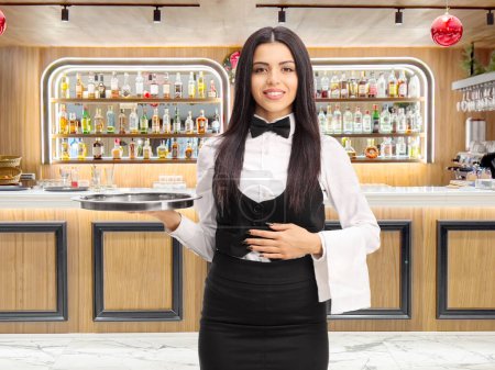 Téléchargez les photos : Jeune serveuse tenant un plateau en métal gris et une serviette blanche devant un bar restaurant - en image libre de droit