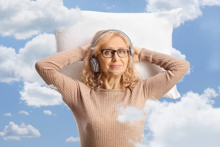 Téléchargez les photos : Femme d'âge moyen avec des écouteurs reposant sur un oreiller parmi les nuages sur un ciel bleu - en image libre de droit