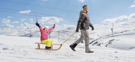 Photo for Full length profile shot of a father pulling a happy little girl on a wooden sled on a snowy mountai - Royalty Free Image