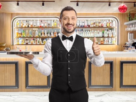 Foto de Camarero sosteniendo una bandeja de plata y mostrando los pulgares delante de un bar - Imagen libre de derechos