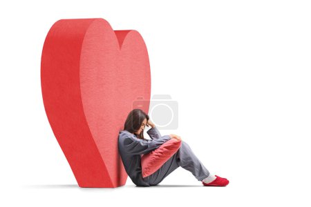 Photo for Sad young woman in pajamas sitting next to a red heart and holding a pillow isolated on white background - Royalty Free Image