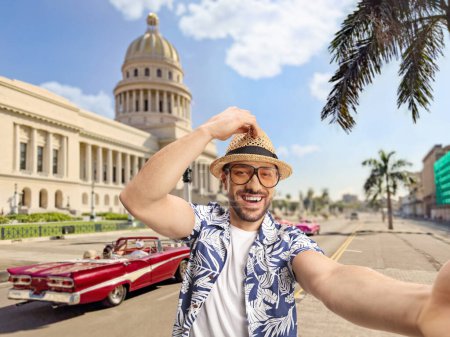 Téléchargez les photos : Joyeux jeune homme avec un chapeau de paille et des lunettes prenant un selfie à La Havane, Cuba, devant El Capitolio - en image libre de droit