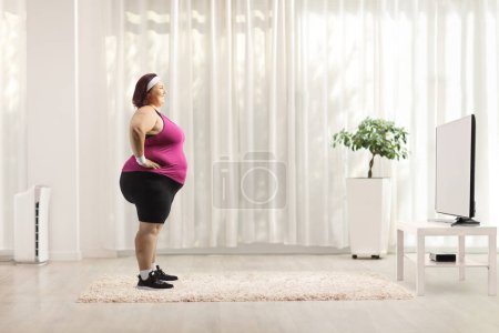 Téléchargez les photos : Plan de profil complet d'une femme de taille plus en vêtements de sport debout devant une télévision à la maison - en image libre de droit
