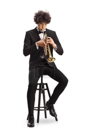 Téléchargez les photos : Élégant jeune homme en costume noir jouant de la trompette et assis sur une chaise isolée sur fond blanc - en image libre de droit