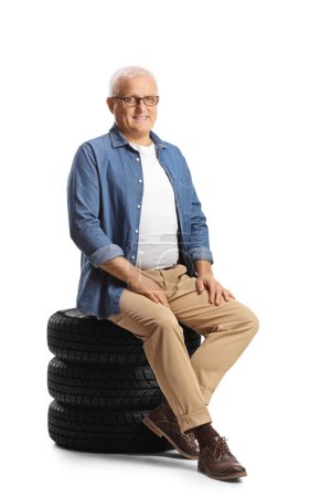 Téléchargez les photos : Homme mûr assis sur un tas de pneus de voiture isolé sur fond blanc - en image libre de droit