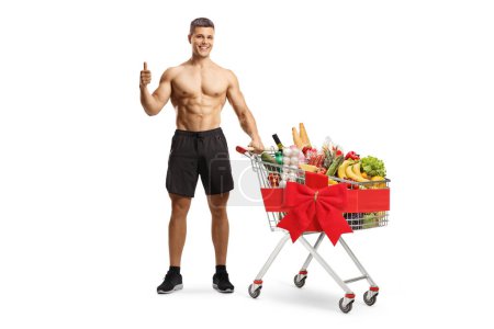 Photo for Shirtless muscular man with a shopping cart full of food and tied with a red ribbon isolated on white background - Royalty Free Image