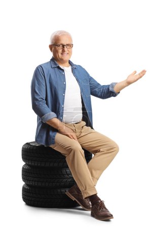 Téléchargez les photos : Homme mûr assis sur un tas de pneus de voiture et pointant isolé sur fond blanc - en image libre de droit