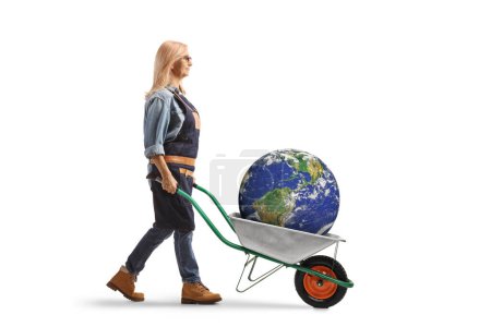 Téléchargez les photos : Vue de profil pleine longueur d'une femme poussant une brouette avec la planète Terre à l'intérieur isolé sur fond blanc - en image libre de droit