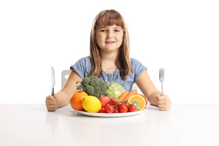 Foto de Niña sentada con un plato de frutas y verduras y sosteniendo un tenedor y un cuchillo aislados sobre fondo blanco - Imagen libre de derechos