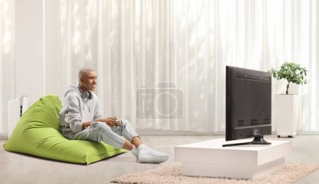 Photo for African american guy sitting on a green beanbag and playing a video game with joystick - Royalty Free Image