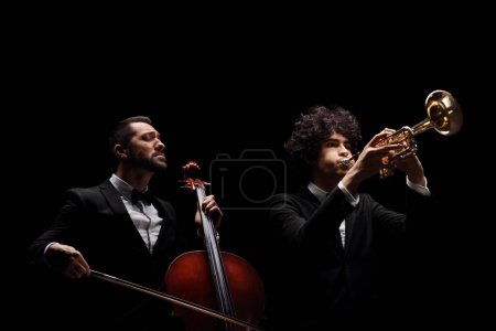 Foto de Músicos masculinos tocando un violonchelo y una trompeta aislados sobre fondo negro - Imagen libre de derechos