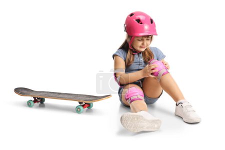 Foto de Chica con monopatín, con rodilleras y casco y sentada en el suelo con la rodilla lesionada aislada sobre fondo blanco - Imagen libre de derechos