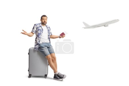 Photo for Confused man holding a passport and sitting on a suitcase waiting for a delayed flight isolated on white background - Royalty Free Image