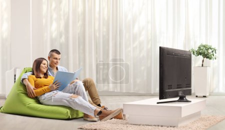 Téléchargez les photos : Jeune couple assis sur un sac à haricots et lisant un livre devant la télévision - en image libre de droit