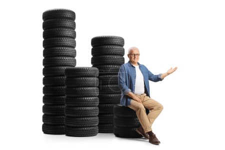 Téléchargez les photos : Homme mûr assis sur un tas de pneus de voiture et montrant à la main isolé sur fond blanc - en image libre de droit