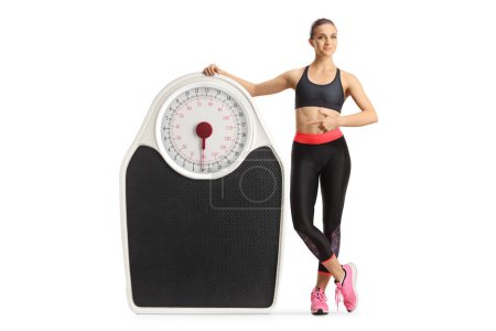 Fit young woman in sportswear pointing at a big weight scale isolated on white background