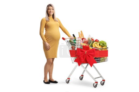 Photo for Pregnant woman standing with a shopping cart full of groceries tied with a red ribbon isolated on white background - Royalty Free Image