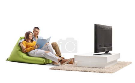 Téléchargez les photos : Jeune couple assis sur un sac à haricots et lisant un livre devant une télévision isolée sur fond blanc - en image libre de droit