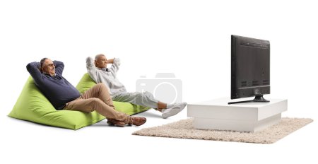 Téléchargez les photos : Homme mûr et homme afro-américain assis sur une chaise de haricot vert et relaxant devant la télévision isolé sur fond blanc - en image libre de droit