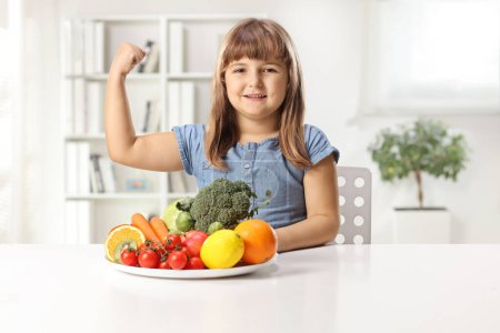 Téléchargez les photos : Petite fille forte assise avec une assiette de fruits et légumes sur une table et montrant le muscle - en image libre de droit