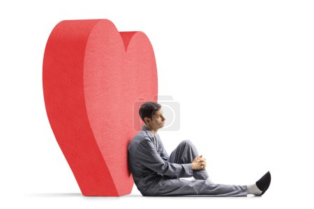 Téléchargez les photos : Triste homme en pyjama assis sur le sol et appuyé sur un cœur rouge isolé sur fond blanc - en image libre de droit