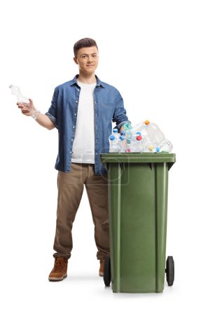 Téléchargez les photos : Jeune homme derrière une poubelle tenant une bouteille en plastique pour recyclage isolé sur fond blanc - en image libre de droit