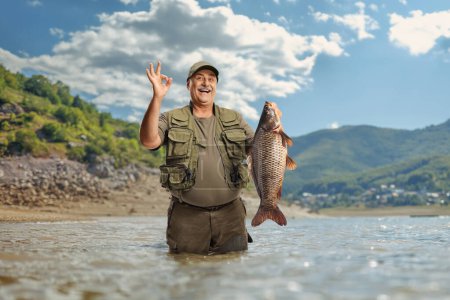 Feliz pescador maduro sosteniendo un pez carpa y haciendo un gesto buena señal dentro de un lago