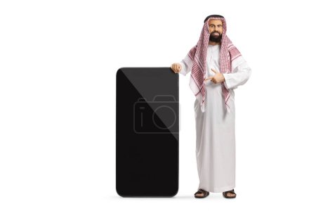 Téléchargez les photos : Saoudien arabe en vêtements ethniques pointant vers un grand téléphone portable avec écran blanc isolé sur fond blanc - en image libre de droit