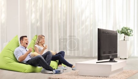 Photo for Joyful man and woman eating popcorn nd sitting on beanbags in front of tv - Royalty Free Image