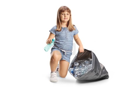 Téléchargez les photos : Petite fille recueillant des bouteilles en plastique dans un sac et souriant à la caméra isolée sur fond blanc - en image libre de droit