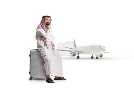 Téléchargez les photos : Homme arabe saoudien assis sur une valise et pensant, annulé ou un concept de vol retardé isolé sur fond blanc - en image libre de droit