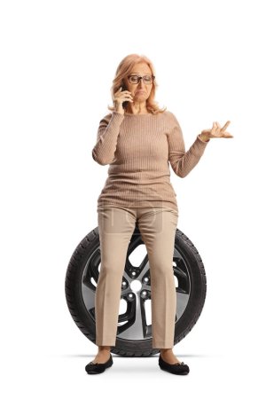 Téléchargez les photos : Photo de profil d'une femme assise sur un pneu de voiture de secours et utilisant un smartphone isolé sur fond blanc - en image libre de droit