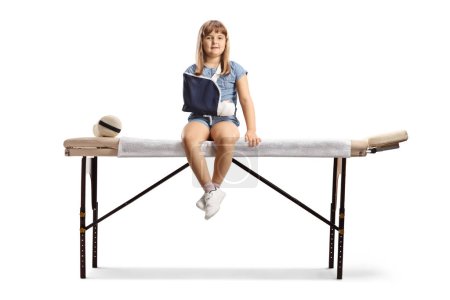 Téléchargez les photos : Portrait complet d'une fille avec un bras cassé portant une fronde et assise sur un lit médical isolé sur fond blanc - en image libre de droit