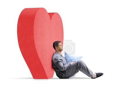 Photo for Pensive young man in pajamas holding a pillow and leaning on a red heart isolated on white background - Royalty Free Image