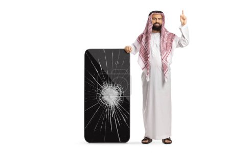 Foto de Hombre árabe saudí con ropa étnica apoyado en un teléfono móvil con la pantalla agrietada y apuntando hacia arriba aislado sobre fondo blanco - Imagen libre de derechos