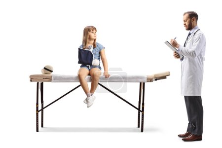 Téléchargez les photos : Médecin écrivant un document et une fille avec un bras cassé portant une fronde et assis sur un lit médical isolé sur fond blanc - en image libre de droit