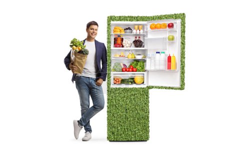 Ganzkörperporträt eines jungen Mannes, der eine Einkaufstasche in der Hand hält und sich an einen energieeffizienten Kühlschrank lehnt, isoliert auf weißem Hintergrund