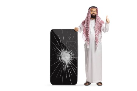 Téléchargez les photos : Saoudien arabe en vêtements ethniques appuyé sur un téléphone portable avec écran fissuré et geste pouces vers le haut isolé sur fond blanc - en image libre de droit