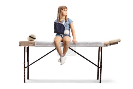 Téléchargez les photos : Petite fille avec un bras cassé portant une fronde et assise sur un lit de thérapie physique isolé sur fond blanc - en image libre de droit