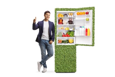 Full length portrait of a young man gesturing thumbs up and leaning on a sustainable fridge isolated on white background