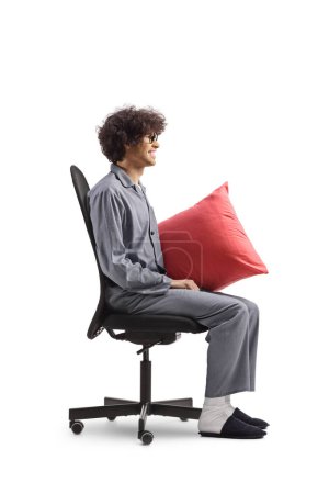 Profilaufnahme eines Mannes im Pyjama, der in einem Bürostuhl sitzt und ein Kissen auf weißem Hintergrund hält.