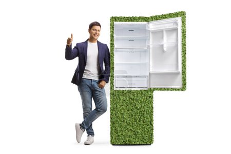Full length portrait of a young man gesturing thumbs upo and leaning on a sustainable fridge isolated on white background