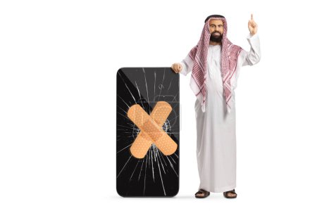 Foto de Hombre árabe saudí con ropa étnica apuntando hacia arriba y de pie junto a un teléfono móvil con la pantalla agrietada aislada sobre fondo blanco - Imagen libre de derechos