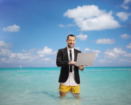 Foto de Empresario en traje y pantalones cortos de baño de pie en el mar y trabajando en un ordenador portátil - Imagen libre de derechos