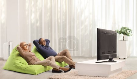 Middle aged man and woman resting on green bean bag armchairs in front of tv 
