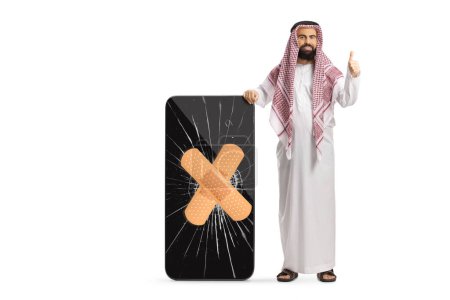 Foto de Hombre árabe saudí con ropa étnica haciendo gestos con los pulgares hacia arriba y de pie junto a un teléfono móvil con la pantalla agrietada aislada sobre fondo blanco - Imagen libre de derechos