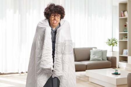 Foto de Joven envuelto en una manta de pie en una sala de estar y sintiéndose enfermo - Imagen libre de derechos