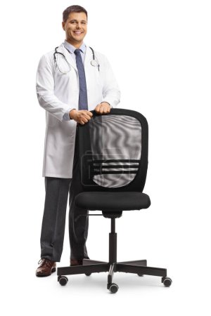 Foto de Médico masculino parado detrás de una silla de oficina y sonriendo aislado sobre fondo blanco - Imagen libre de derechos