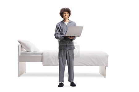 Photo for Young man in pajamas standing in front of a bed and working on a laptop computer isolated on white background - Royalty Free Image