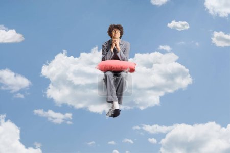 Téléchargez les photos : Jeune homme en pyjama assis sur un nuage dans le ciel et pensant - en image libre de droit